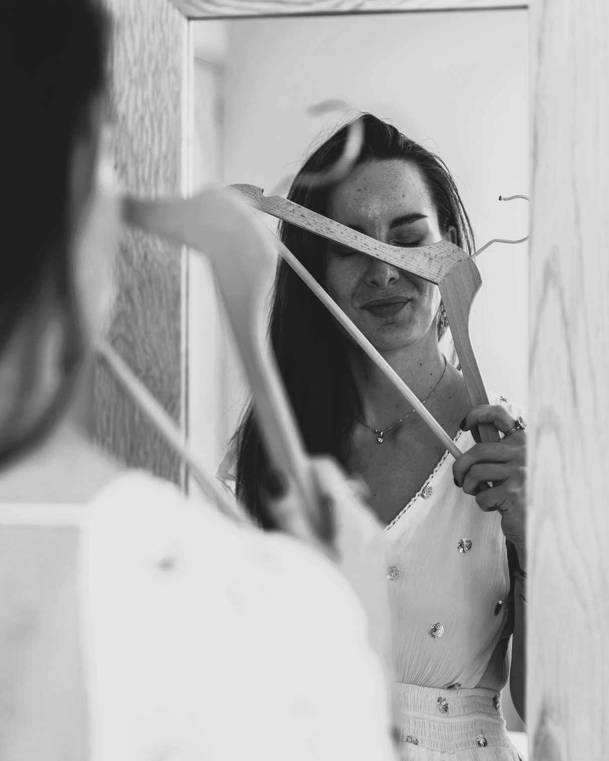 Femme en robe blanche se regardant dans un miroir, tenant des cintres devant son visage, expression douce et introspective.