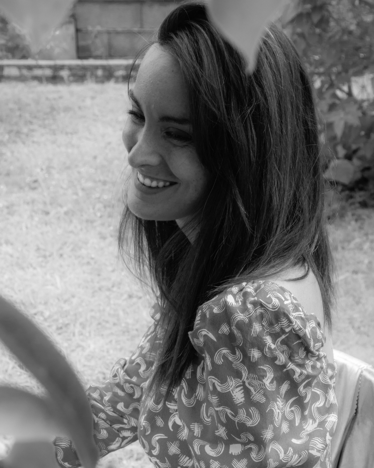 Femme souriante en extérieur, vêtue d’une robe à motifs, photographiée en noir et blanc